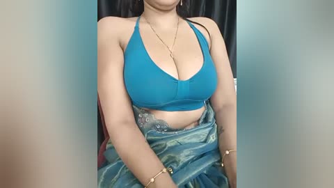 Media: Video of a woman with medium skin tone wearing a teal halter top, revealing cleavage, and a blue sari with a gold bling. She has long, dark hair and is seated against a teal backdrop.