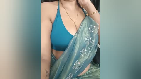 A video of a woman wearing a turquoise halter top and a green sari with silver embroidery, against a plain background. She has a gold necklace and bracelet, and a small tattoo on her wrist.