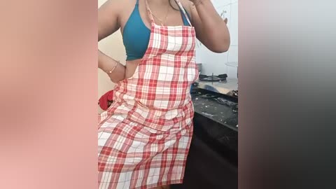 Media: A video of a person with medium brown skin, wearing a teal sports bra and a red plaid apron, standing in a kitchen with cluttered counters.