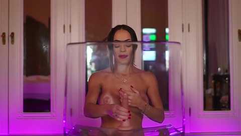 Media: Video of a topless woman with long dark hair, holding a transparent, cylindrical object over her breasts, indoors with purple lighting, white-framed windows, and modern decor in the background.