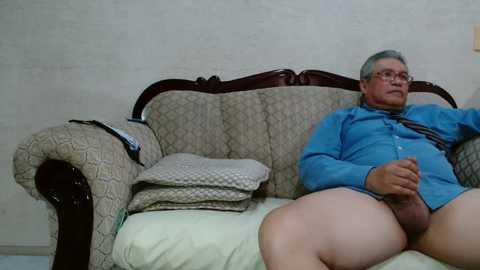 Media: A video of an elderly man with glasses, wearing a blue shirt and no pants, sitting on a beige-patterned couch, surrounded by pillows.