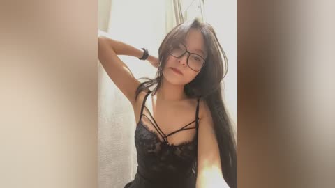 Video of an Asian woman with long black hair, wearing black lace lingerie and glasses, posing seductively in a dimly lit room with a white wall and window.