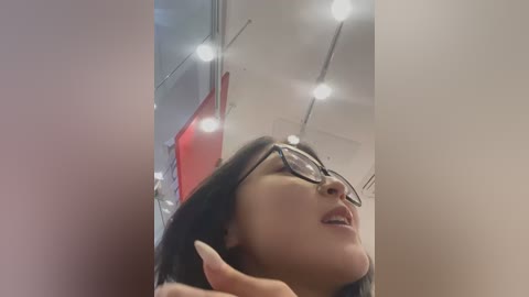 Media: A video of a woman with straight black hair, wearing glasses, and a light-colored top, captured from a low angle, with a blurred red background and bright ceiling lights.