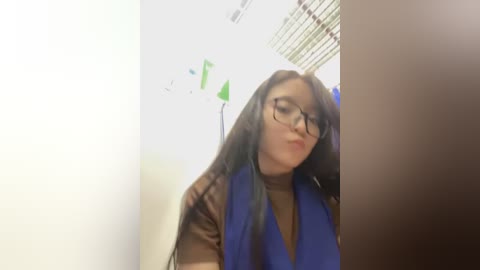 Media: Video of a young woman with long black hair, wearing glasses, a blue vest, and a brown top, standing in a narrow hallway with green decorations and fluorescent lighting.