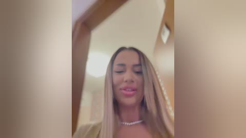 Media: Video of a young woman with long, straight, blonde hair, wearing a pearl necklace, and a light-colored top, looking down, with blurred background of a room with beige walls and a ceiling light.