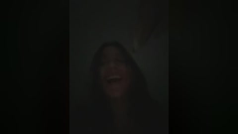 Media: A blurry video of a person with long dark hair and a smile, standing in a dimly lit room, creating a mysterious and slightly eerie atmosphere.