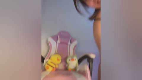 Media: A blurred video shows a person with short brown hair, partially visible, sitting in a pink and white gaming chair. Two yellow and green plush toys are placed on the chair.