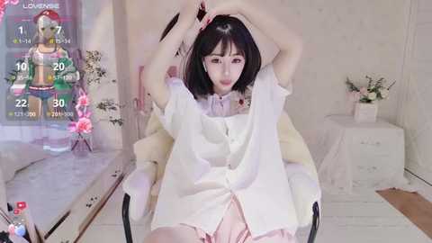 Media: Video of a young Asian woman with straight black hair, wearing a white blouse and pink skirt, sitting in a white chair, stretching arms. Background features a floral arrangement and a video game overlay.