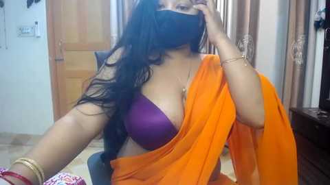 Media: Video of a woman with long black hair, wearing a purple bra, orange sari, and black face mask, sitting on a chair in a brightly lit room with beige curtains and wooden doors.