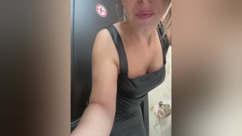 Media: Video of a light-skinned woman with blonde hair, wearing a black dress, taking a selfie in a bathroom with a tiled background and a visible sink.