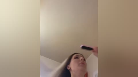 A video of a woman with long brown hair, wearing a white top, holding a black remote control, taken from a low angle, showing the ceiling and upper body.