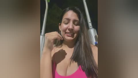 Video of a smiling Latina woman with long, straight dark hair, wearing a low-cut pink tank top, standing in a sunlit park, holding a metal pole.