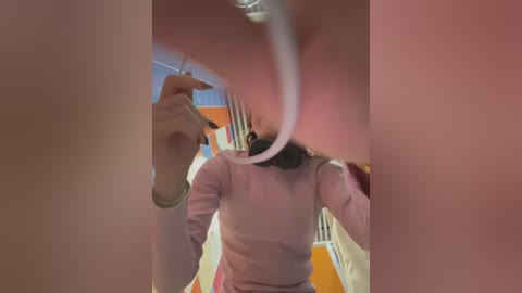 Video of a woman taking a selfie in a bathroom mirror, with a visible reflection of her hand holding the phone. She's wearing a pink sweater and has shoulder-length dark hair.