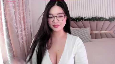 Media: A video of a young Asian woman with long black hair, wearing large round glasses, a low-cut white blouse, and a seductive expression. She stands in a stylish, pink-hued bedroom with a tufted headboard and floral curtains.