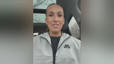 Media: Video of a young woman with light brown skin and straight hair tied back, wearing a light gray Nike jacket with a black Nike logo, sitting in a car with bare trees outside.