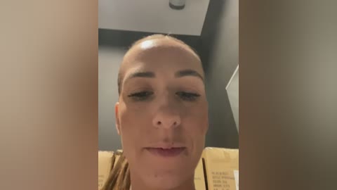 Media: A video of a bald Caucasian woman with light skin, wearing makeup, including dark eyeliner and mascara, and a brown top, with a blurred background featuring a white ceiling and beige wall.