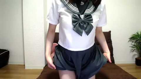 Media: A video of an Asian woman wearing a Japanese schoolgirl outfit, including a white blouse with a large green bow, and a short pleated skirt. She stands in a room with a white wall, brown couch, and potted plant.