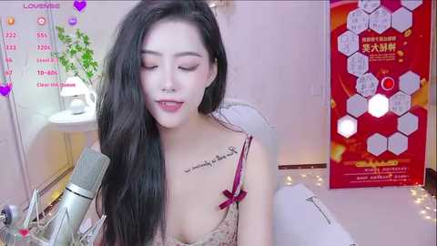 Media: Video of an East Asian woman with long black hair, fair skin, wearing a pink lace bralette, smiling, with a tattoo on her chest, surrounded by recording equipment and promotional materials in a soft, pastel-toned room.