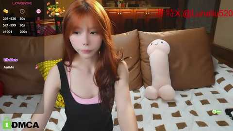 Media: A video of a young Asian woman with long, straight red hair, wearing a pink tank top under a black dress, sitting on a brown leather couch. The background shows a kitchen with wooden cabinets and a white patterned blanket.