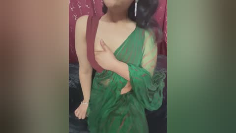 Media: Video of an Asian woman with medium skin tone, wearing a sheer green blouse, partially covering her chest. She's standing in front of a red, patterned curtain.