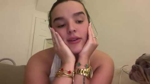 Media: Video of a young woman with fair skin and dark hair, wearing gold bracelets, resting her head on her hands, looking pensive in a beige room.