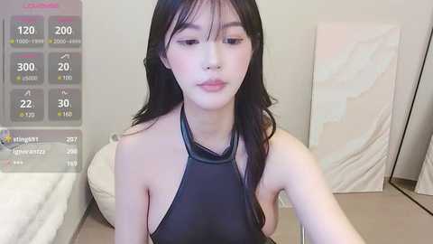 Media: Video of an East Asian woman with long black hair, wearing a black halter top, sitting on a beige couch. Background features a white abstract painting and a partially visible bed.