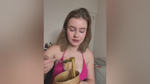 Video of a young, light-skinned woman with shoulder-length brown hair, wearing a pink halter top, eating noodles from a bowl. Background shows a messy room with a bed and scattered objects.