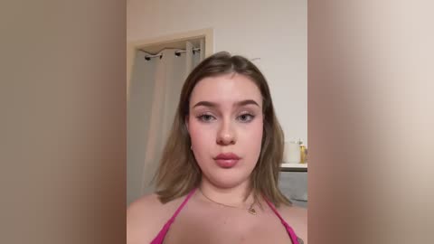 Video of a young Caucasian woman with light skin, shoulder-length brown hair, and blue eyes, wearing a pink halter top. She has a neutral expression and is standing in a simple, beige-colored bathroom with a shower curtain in the background.