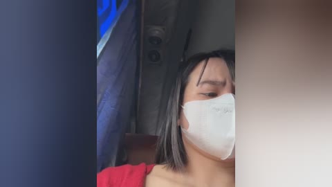 Media: A video of a woman with straight black hair, wearing a white surgical mask, red shirt, and looking through a narrow gap.