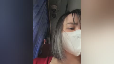 Media: Video of an East Asian woman with short black hair, wearing a white mask, a red shirt, and blue curtains, taken indoors.