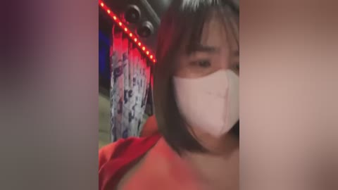 A video of a woman with straight black hair, wearing a face mask, and a red shirt, standing in a dimly lit room with red LED lights and a hanging white patterned tapestry.