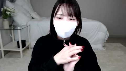 Media: Video of a young Asian woman with long black hair, wearing a white face mask, black top, and red nail polish, sitting on a white rug in a minimalist, grey-toned bedroom.