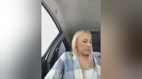 Media: Video of a middle-aged woman with long blonde hair, wearing a plaid shirt, resting her head on a car seat. The car interior is dimly lit with a blurry, out-of-focus background.