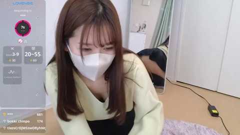 Video of an Asian woman with long brown hair and a white mask, kneeling in a bedroom, wearing a yellow top and black shorts, with a virtual reality headset on.