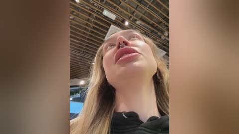 Media: Video of a young Caucasian woman with long, light brown hair, wearing a black top, looking upward with a slight smile. The background features a wooden ceiling and visible lights.