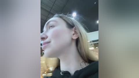 Media: A video of a young woman with fair skin and straight blonde hair, wearing a black top, captured from a low angle, showing her profile. The background is a blurred indoor setting with soft lighting and a modern ceiling.