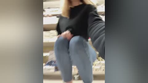 A blurry video of a person sitting on a bench, wearing a black sweater and blue jeans, with a blurred background showing shelves and a store setting.