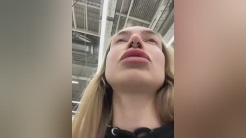 Video of a young woman with fair skin, long blonde hair, and full lips, taken from a low angle, showing her from below the nose to the top of her head. Background is an industrial ceiling with metal beams.