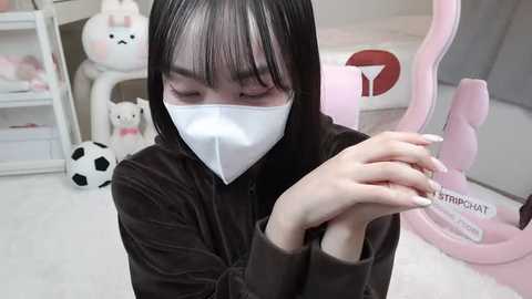 Media: A video of an Asian woman with straight black hair and pale skin, wearing a white mask, black hoodie, and white gloves, carefully holding a small plastic animal in a pastel-colored room with plush toys and a pink gaming chair.