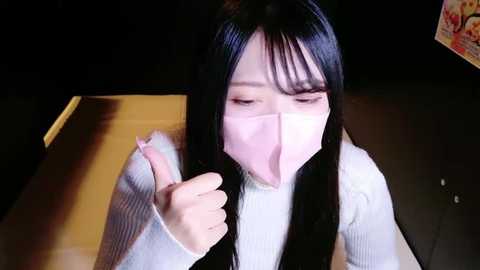 Media: Video of a young East Asian woman with long black hair, wearing a white face mask and a light grey sweater, giving a thumbs-up in a dimly lit room.