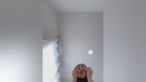 Media: A video of a young woman with long black hair, wearing a black top, standing in a minimalist, white-walled room with a small circular light fixture above her.