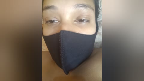 Media: A video of a person with medium brown skin, wearing a black surgical mask covering the nose and mouth, and a black headband. The background is blurred and neutral-colored.