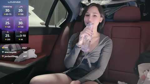 Media: Video of a young Asian woman with light skin, sitting in a car, wearing a gray top and black skirt, drinking from a clear plastic bottle, with a digital screen displaying location and temperature information.