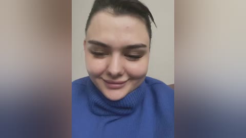 Media: Video of a young woman with fair skin and dark hair, wearing a blue turtleneck sweater, smiling and closing her eyes. Background is blurred.