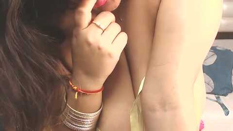 Media: A close-up video of a woman with long dark hair and fair skin, wearing a red bindi and gold bracelets, gently touching her cheek. Her yellow garment is partially visible.