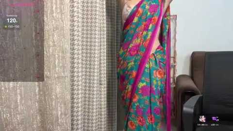 Media: A video of a woman wearing a vibrant, floral-patterned sari, partially covering her chest with a pink blouse. The background features textured wallpaper and a brown leather chair.