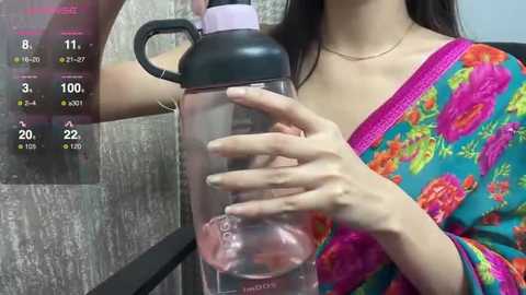 Media: Video of a woman with long black hair, wearing a vibrant floral sari, filling a large water bottle with a black cap. The background features a textured gray wall and a digital display showing water consumption stats.