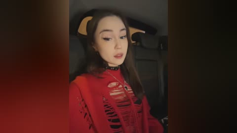 Video of a young Asian woman with fair skin, wearing a red, torn, and distressed t-shirt with exposed skin, and devil horns headband. She has long, straight brown hair and is seated in a dimly lit car.