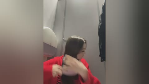 Media: Video of a woman with long, straight black hair, wearing a bright red sweater, combing her hair in a narrow, dimly-lit bathroom stall.