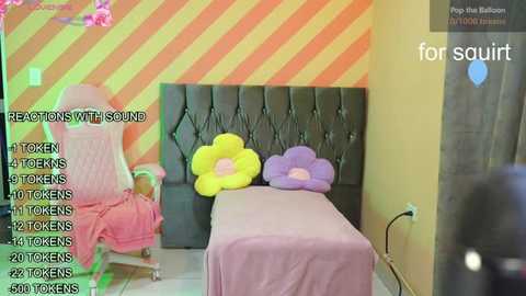 Video of a whimsical bedroom with a pink gaming chair, a tufted black headboard, and oversized, plush yellow and purple flower pillows, set against a striped orange and yellow wall.